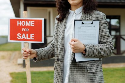Real estate agent holding "Home for Sale" sign and clipboard for first-time homebuyers.